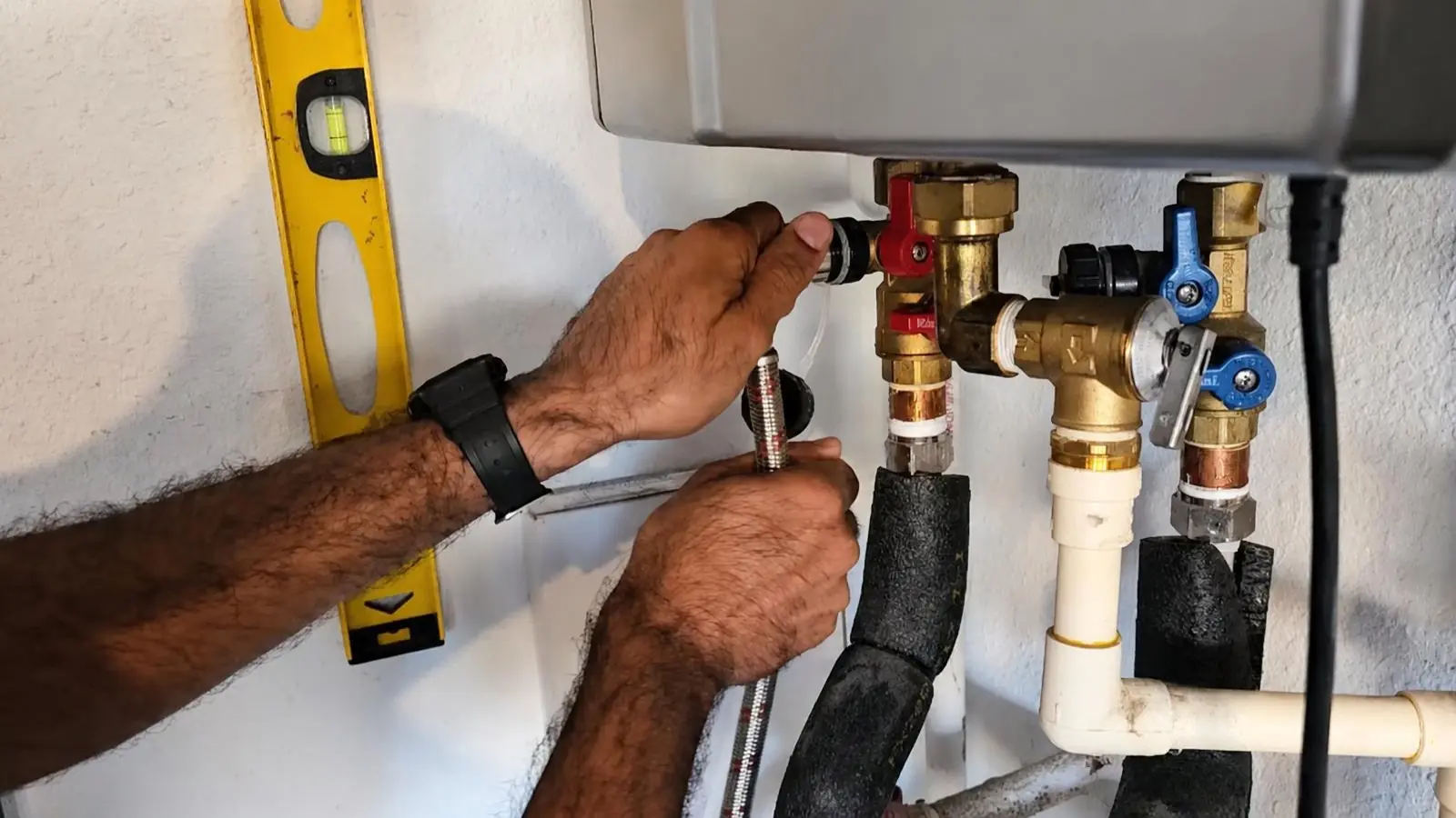 Hands connecting service valves and a flexible hose under a wall-mounted tankless water heater